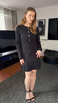 Svart kjole med lange ermer, knappelukking foran, volangkant nederst og tettsittende silhuett. Modellen har på seg svarte remmesandaler med hæl, og står på et teksturert teppe.