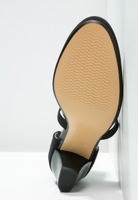 Scarpa nera con tacco alto, con parte superiore nera testurizzata e suola in gomma color beige chiaro con motivo a onda, inclinata su uno sfondo bianco.