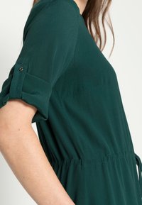 Vestido verde-azulado feito de tecido leve, com mangas roladas curtas, cintura elástica com cordão e detalhes de costura vertical.