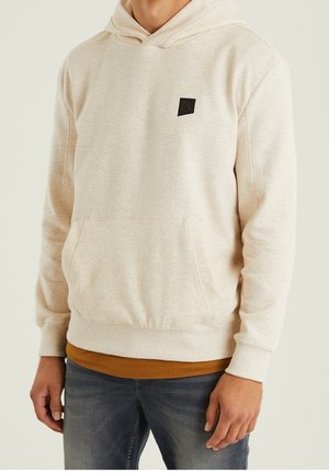 Mann trägt einen strukturierten, elfenbeinfarbenen Kapuzenpullover mit einer Fronttasche über einem braunen Hemd und verblassten blauen Jeans, steht vor einem einfarbigen Hintergrund.