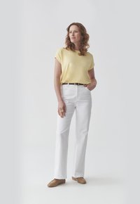 Geel top met korte mouwen, gecombineerd met witte rechte broek en lichtbruine platte schoenen. Model draagt een zwarte riem en een ring.