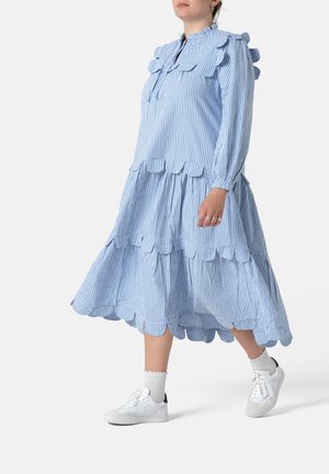 Femme marchant dans une robe vichy bleue et blanche avec des bords festonnés, des chaussettes blanches et des baskets blanches sur un fond uni.