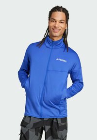 Sininen vetoketjullinen takki, jossa on korkea kaulus, valmistettu sileästä kankaasta. Takissa on rintatasku ja valkoinen "Terrex"-logo.