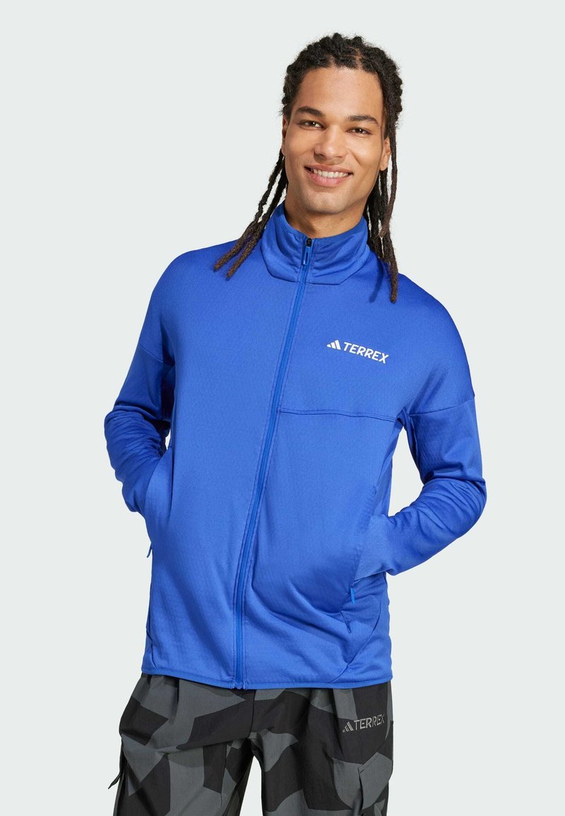 Sininen vetoketjullinen takki, jossa on korkea kaulus, valmistettu sileästä kankaasta. Takissa on rintatasku ja valkoinen "Terrex"-logo.