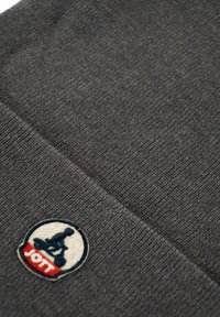 Donkergrijs gebreid stof met een ronde patch waarop een blauwe zittende figuur en rode tekst "JOTT" op een witte achtergrond.