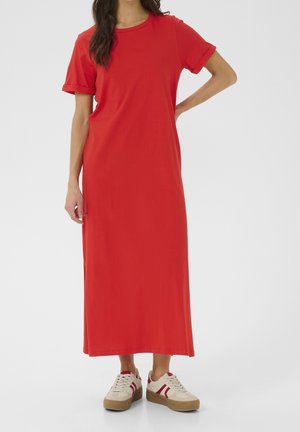 Frau trägt ein rotes Midi-Kleid mit kurzen Ärmeln und weiße Turnschuhe mit roten Streifen, steht vor einem schlichten hellen Hintergrund.