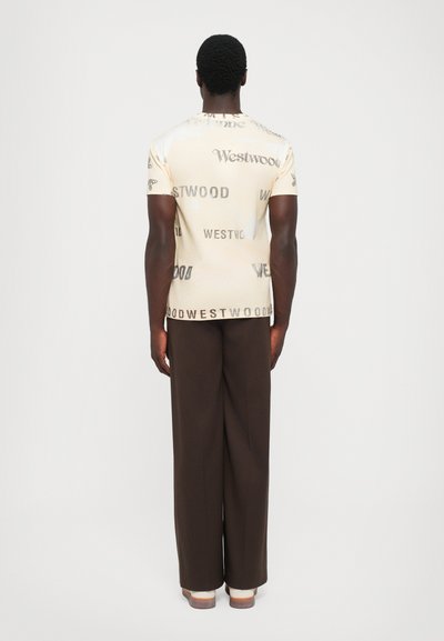 Bărbat în picioare, întors cu spatele, purtând un tricou bej cu mai multe logo-uri "Westwood" și pantaloni largi maro închis, cu pantofi albi.