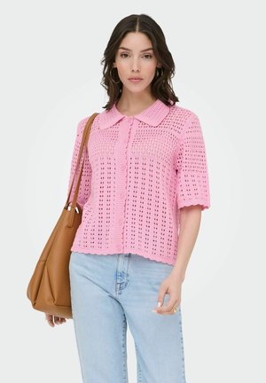 Kobieta w różowym, szydełkowanym kardiganie z krótkim rękawem, jasnoniebieskich dżinsach, złotych kolczykach kołach, niesie brązową skórzaną torebkę na ramię.