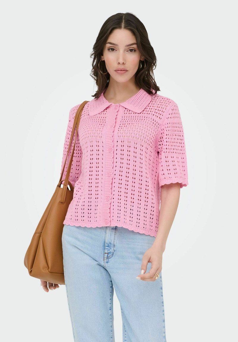 Žena nosi roza kukičani kardigan s kratkim rukavima, svijetloplave traperice, zlatne naušnice u obliku obruča i nosi smeđu kožnu torbu preko ramena.