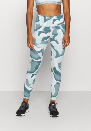 Leggings à motif camouflage bleu clair et vert, fabriqués en matériau extensible, avec une taille haute et une coupe ajustée, associés à des baskets noires.
