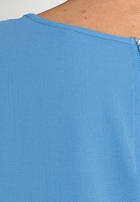 Modrý top s hladkým povrchem, zakulaceným výstřihem a diskrétním zipem na zadní straně. Materiál vypadá lehký a měkký.