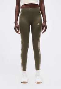 Άτομο που φοράει λαδί κολάν Adidas με μπεζ ρίγες στα πλαϊνά, λευκές κάλτσες και λευκά αθλητικά παπούτσια, στέκεται μπροστά σε ένα απλό φόντο.