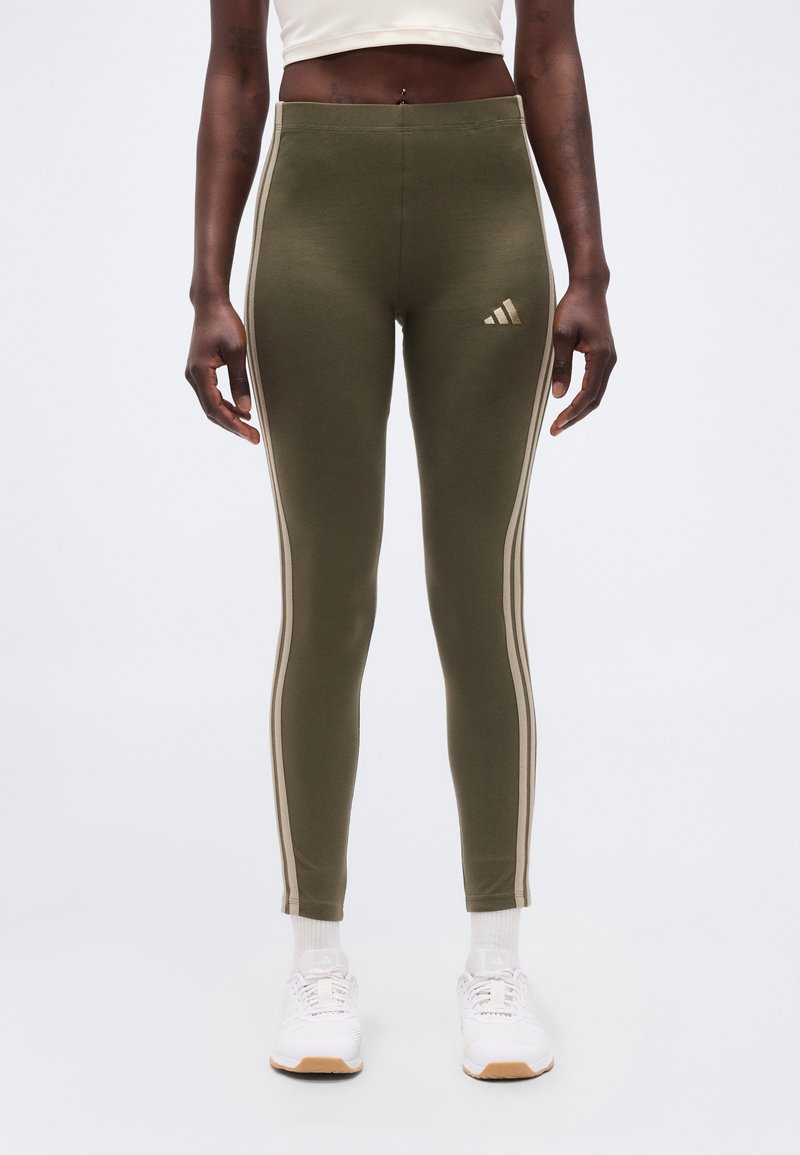 Άτομο που φοράει λαδί κολάν Adidas με μπεζ ρίγες στα πλαϊνά, λευκές κάλτσες και λευκά αθλητικά παπούτσια, στέκεται μπροστά σε ένα απλό φόντο.