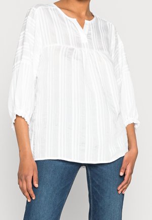 Woman wearing a loose white striped blouse with 3/4 sleeves and gathered cuffs, paired with blue jeans against a neutral background.