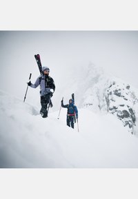 Two skiers ascending a snowy slope, wearing purple and blue jackets, carrying black skis, with gray rocky mountains in the background.
