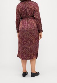 Lungo vestito bordeaux con motivo paisley, vita con cintura e maniche con bottoni. Indossato con scarpe nere slip-on.