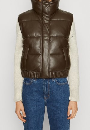 Personne portant un gilet en cuir matelassé marron foncé sur un pull en tricot crème et un jean en denim bleu, debout devant un fond uni.