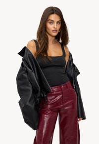 Vrouw met lang bruin haar, gekleed in een zwarte tanktop, een oversized zwart leren jasje dat van haar schouders hangt, en hoge rode krokodillenprintbroek.