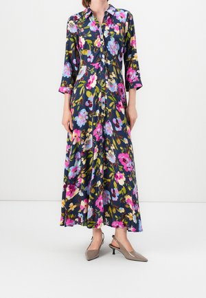 Kobieta ubrana w długą granatową sukienkę w kwiaty w kolorach różowym, fioletowym i żółtym, z przodu zapinaną na guziki, z rękawami do połowy przedramienia i beżowymi czółenkami na niskim obcasie.