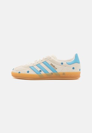 Biały sneaker Adidas Gazelle z jasnoniebieskimi paskami, gumową podeszwą oraz małymi niebieskimi wzorami kwiatowymi na zamszowej cholewce i pętli na pięcie.