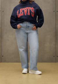 Osoba w granatowej bluzie Levi's, jasnoniebieskich spodniach typu high-waist i białych trampkach, stojąca na tle szarej ściany.