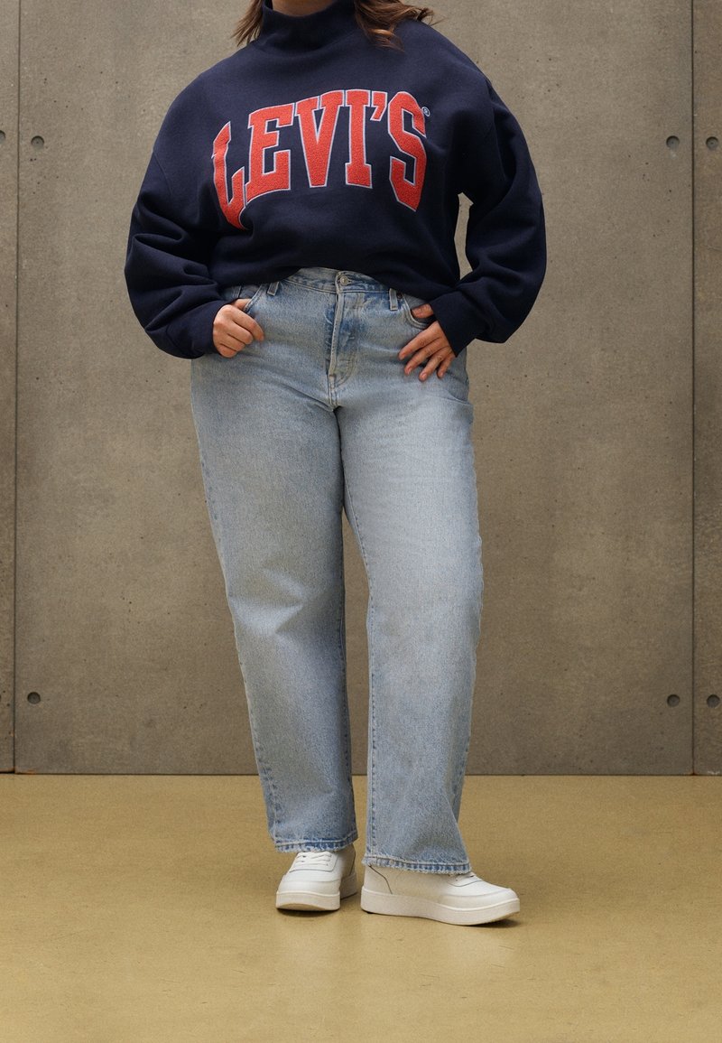 Osoba w granatowej bluzie Levi's, jasnoniebieskich spodniach typu high-waist i białych trampkach, stojąca na tle szarej ściany.
