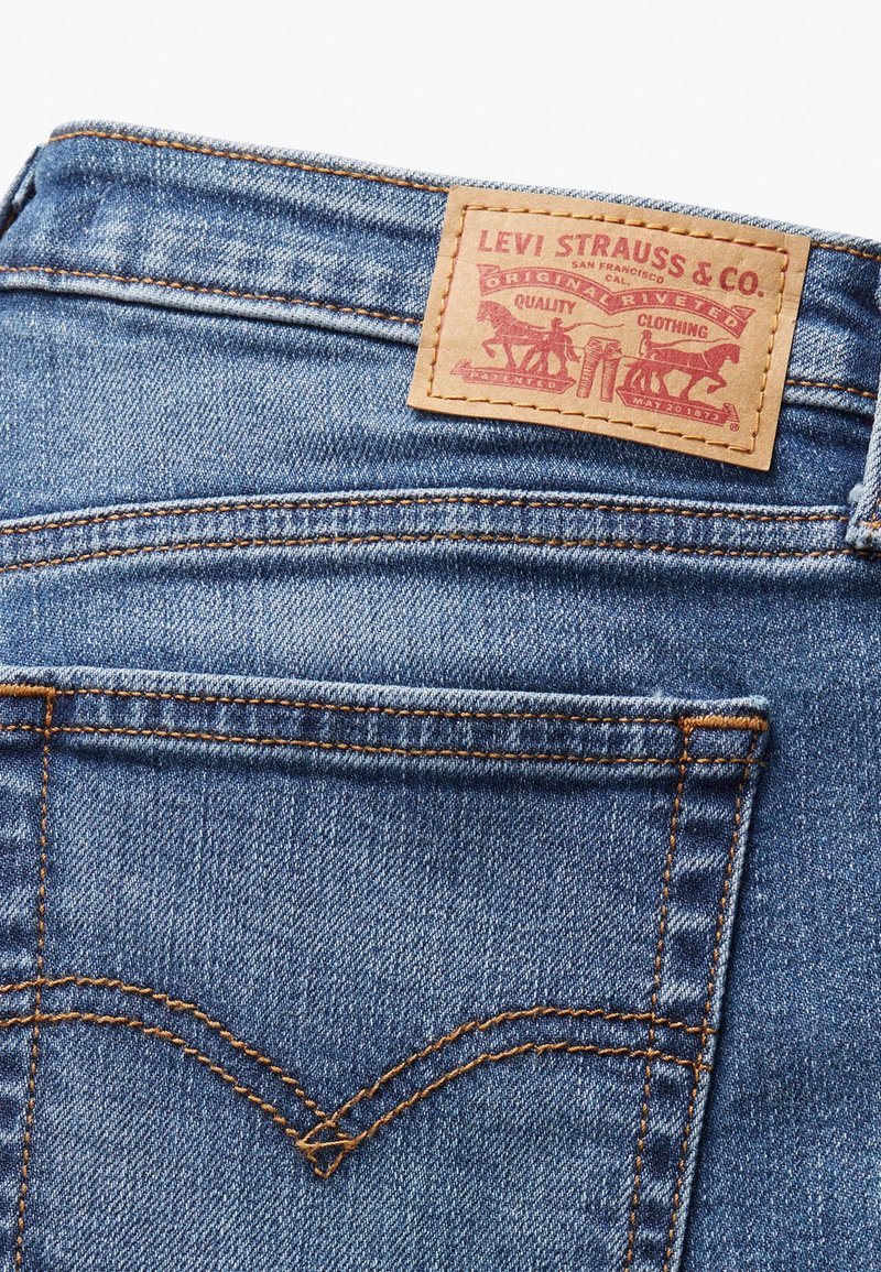 Jeans de mezclilla azul con un parche de cuero con letras rojas. Las costuras son de un naranja contrastante, destacando los detalles de los bolsillos y las costuras.