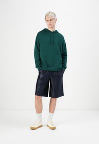 Felpa verde con tasca frontale, abbinata a pantaloni corti blu scuro e sneakers di colore chiaro. Tessuto semplice, vestibilità casual e coulisse regolabile.