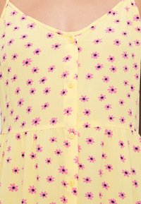 Κίτρινο φόρεμα με λουλουδάτο μοτίβο, που διαθέτει μικρά ροζ λουλούδια με μαύρα κέντρα, λεπτές τιράντες και κίτρινα κουμπιά στο εμπρός μέρος.