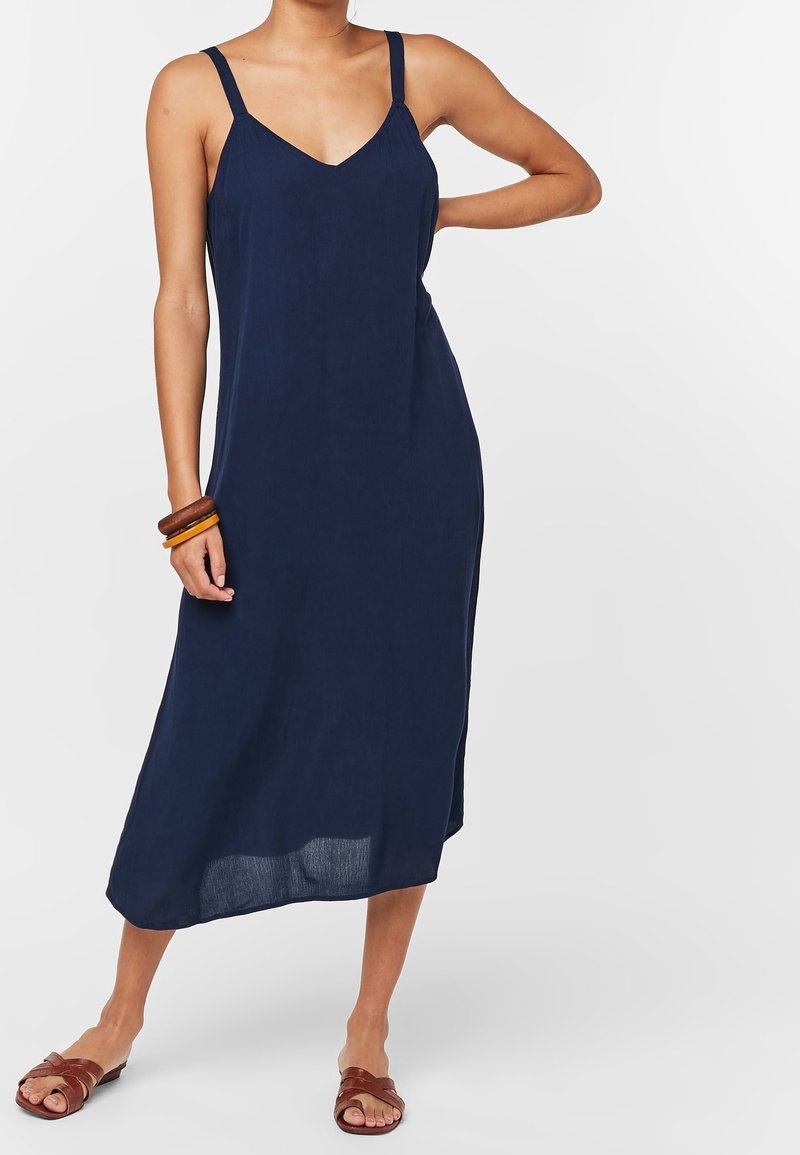Femme portant une robe midi sans manches bleu marine à fines bretelles, des sandales marron à enfiler et des bracelets en bois empilés au poignet gauche.