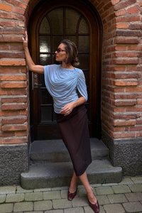 Blouse drapée bleu clair avec un motif à pois, associée à une longue jupe marron foncé. Le mannequin porte des escarpins pointus bordeaux. Mur en briques en arrière-plan.