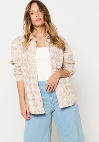 Light beige snake print button-up shirt with long sleeves, front pockets, and light blue wide-leg jeans. Smooth texture and relaxed fit.