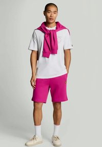 Vit t-shirt med rund hals, rosa shorts och en rosa tröja som hänger runt axlarna. Har en liten logotyp på shortsen.