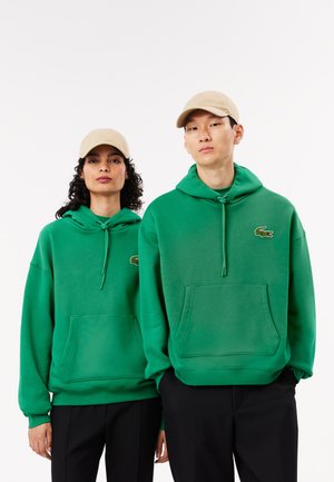 Felpe verdi con cappuccio e tasche a marsupio, con logo ricamato. Indossate con cappelli beige, entrambi i modelli hanno una vestibilità rilassata.