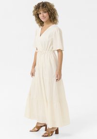 Long, cream-colored maxi dress with a wrap design, short sleeves, elastic waist, tiered skirt, and textured fabric. Paired with brown heeled sandals.