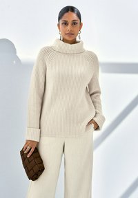 Beige gerippter Pullover mit hohem Kragen und langen Ärmeln, kombiniert mit hellbeigen weiten Hosen. Hält eine braune gesteppten Clutch.