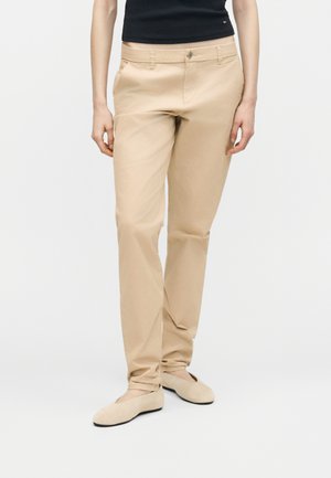 Beżowe spodnie o prostych nogawkach noszone z beżowymi balerinami i czarnym bezrękawnikiem, pokazane od talii do stóp na białym tle.