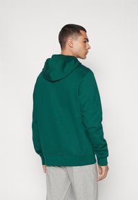 Groene hoodie van zacht materiaal, met een trekkoord voor de capuchon en geribde manchetten. De achterkant is eenvoudig met een losse pasvorm, gecombineerd met grijze joggingbroeken.