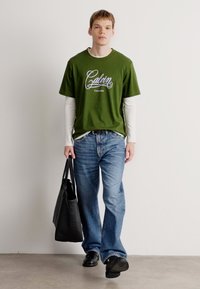 Young man walking, wearing green Calvin Klein T-shirt over white long sleeves, blue jeans, black shoes, carrying a black tote bag.