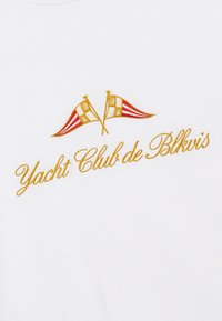 Drapeaux de yacht rouges et blancs brodés en croix au-dessus d’un texte en cursive dorée indiquant « Yacht Club de Blkvis » sur un tissu blanc.