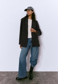 Blazer de cuadros gris cruzado sobre una blusa beige, combinado con jeans de pierna ancha azules y botas negras hasta la rodilla, contra un fondo liso.