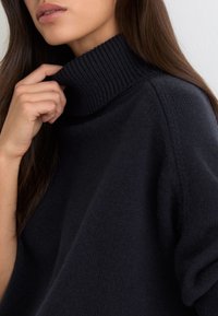Femme ajustant le col côtelé d'un pull à col roulé noir, montrant des lèvres et de longs cheveux bruns sur un fond uni.