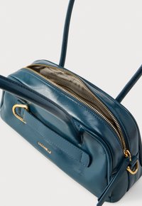 Μπλε δερμάτινο τσάντα crossbody με χρυστό φερμουάρ, μπροστινή λαβή και ρυθμιζόμενο ιμάντα, μερικώς ανοιχτή που δείχνει μπεζ υφασμάτινη επένδυση.