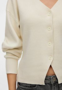Crème-kleurige gebuttonde cardigan gedragen met zwarte gewassen jeans, met een tussenruimte tussen de cardigan en de broekband van de jeans.