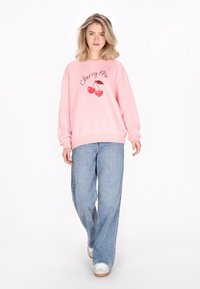 Felpa rosa chiaro con testo "Cherry Me" e grafica di ciliegia; abbinata a jeans blu larghi e sneakers bianche; design rilassato.