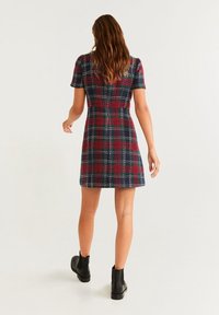 Kurzärmliges, knielanges Kleid im Karomuster aus Marineblau, Rot und Grün mit taillierter Taille, ausgestelltem Saum und glatter Textur. Schwarze Knöchelstiefel.
