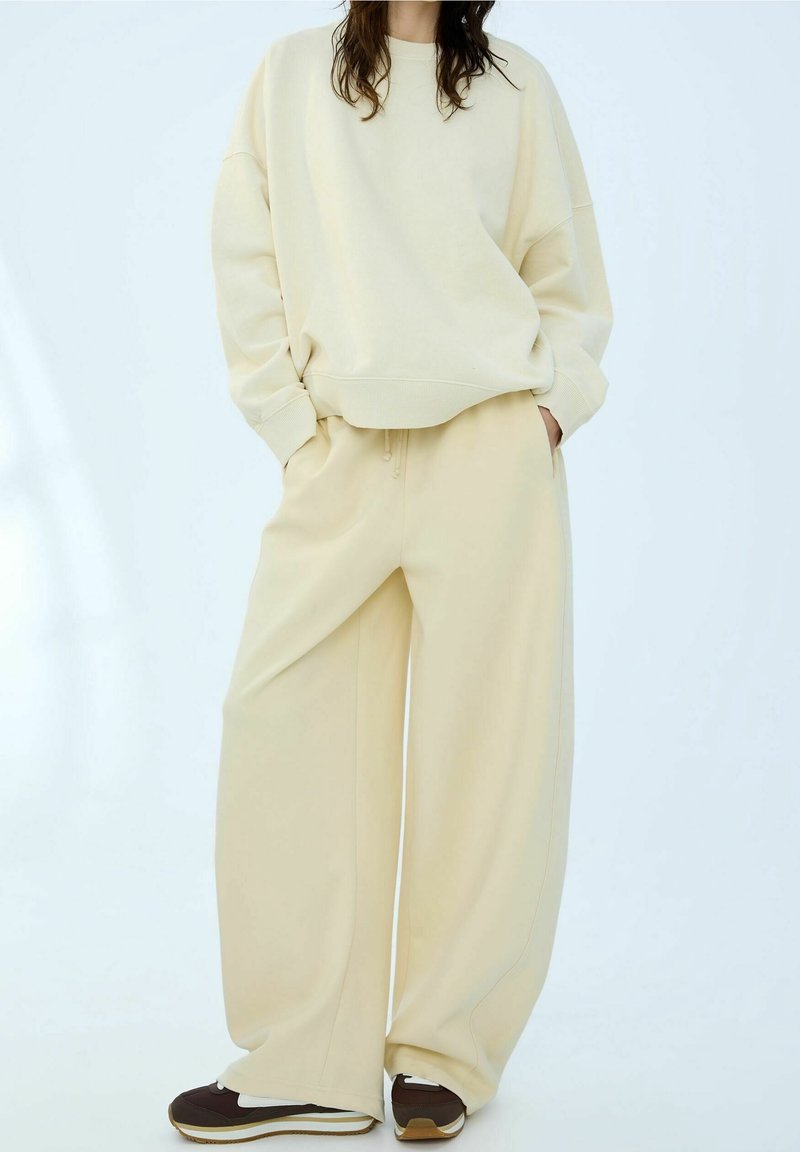 Personne portant un sweat-shirt ample couleur crème et un pantalon large assorti, debout les mains dans les poches et des baskets marron sur fond blanc.