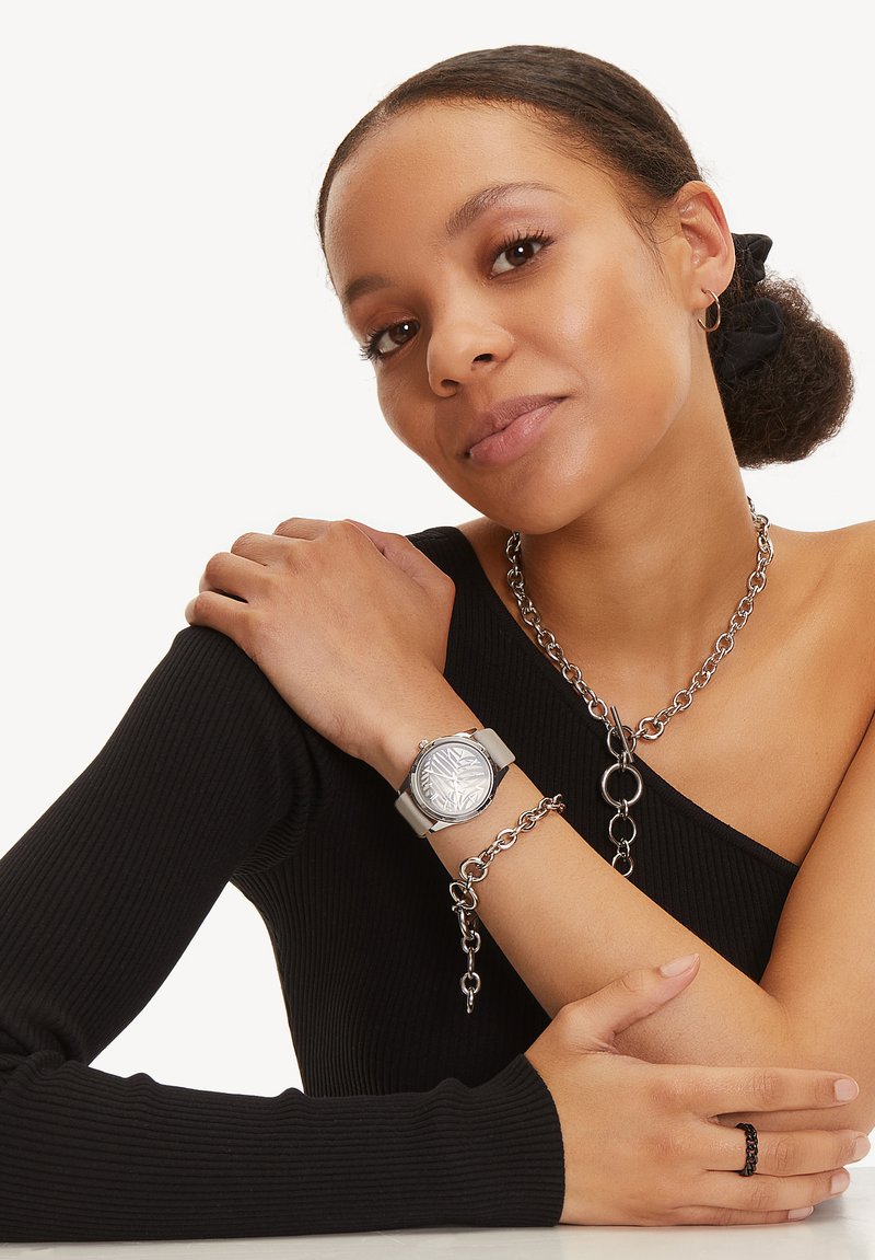 Zilveren polshorloge met een gestructureerde wijzerplaat en een beige band, gecombineerd met een chunky zilveren ketting en een zwarte ring op een gestructureerd zwart top.