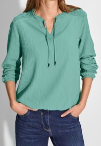 Blouse turquoise froissée avec col en V, cordon de serrage ajustable et manches longues. Portée avec un jean bleu foncé, le tissu présente une finition texturée.
