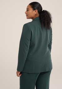 Tmavě zelený upravený blazer z pohledu zezadu, s čistými liniemi, hladkou texturou a strukturovaným siluetou. Jednoduchý design klopy.